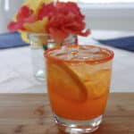 An orange drink with an orange slice in the glass and orange sugar on the rim next to flowers.