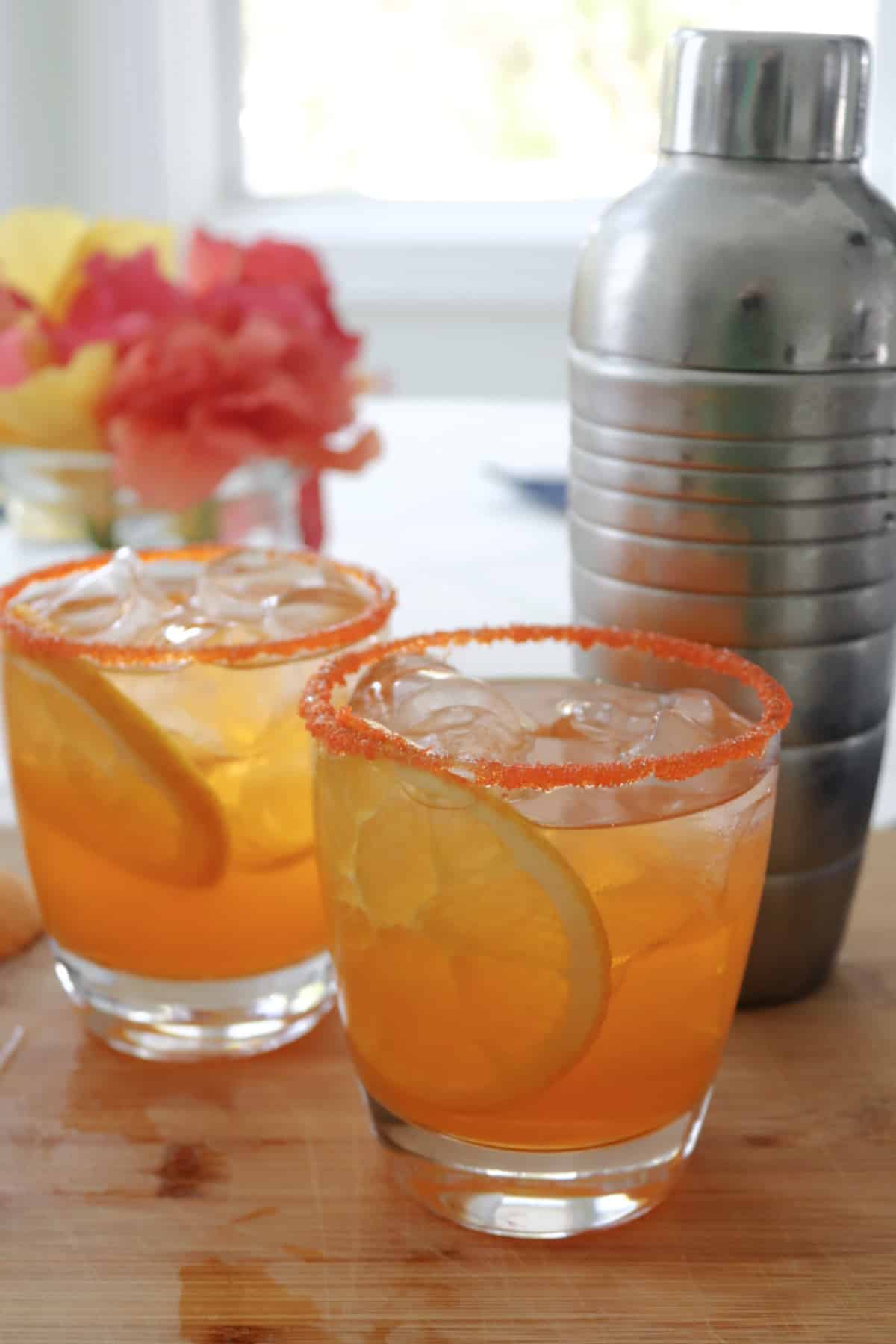 Two sparkling orange margaritas on a board next to a cocktail shaker with flowers in the background.