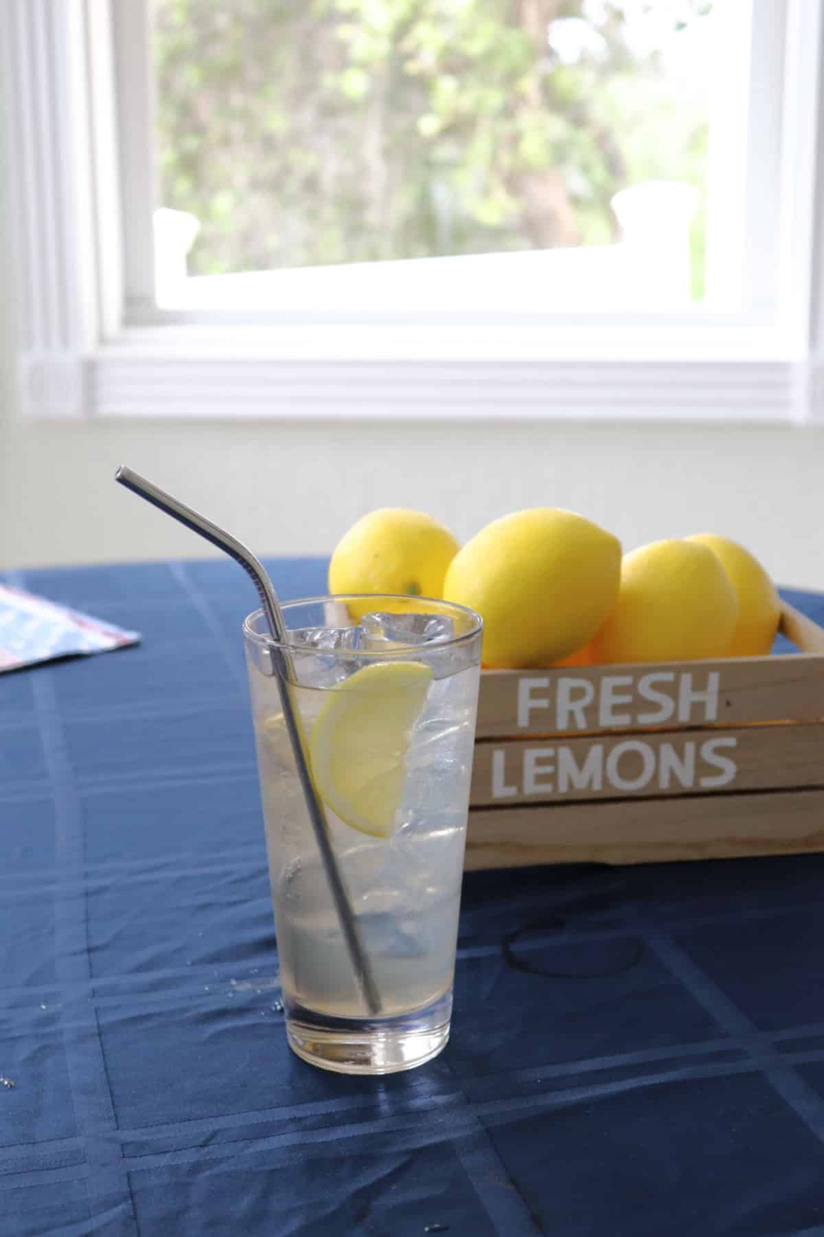 Lemonade Tequila Cocktail in a collins glass next to a crate of lemons on a blue tablecloth.