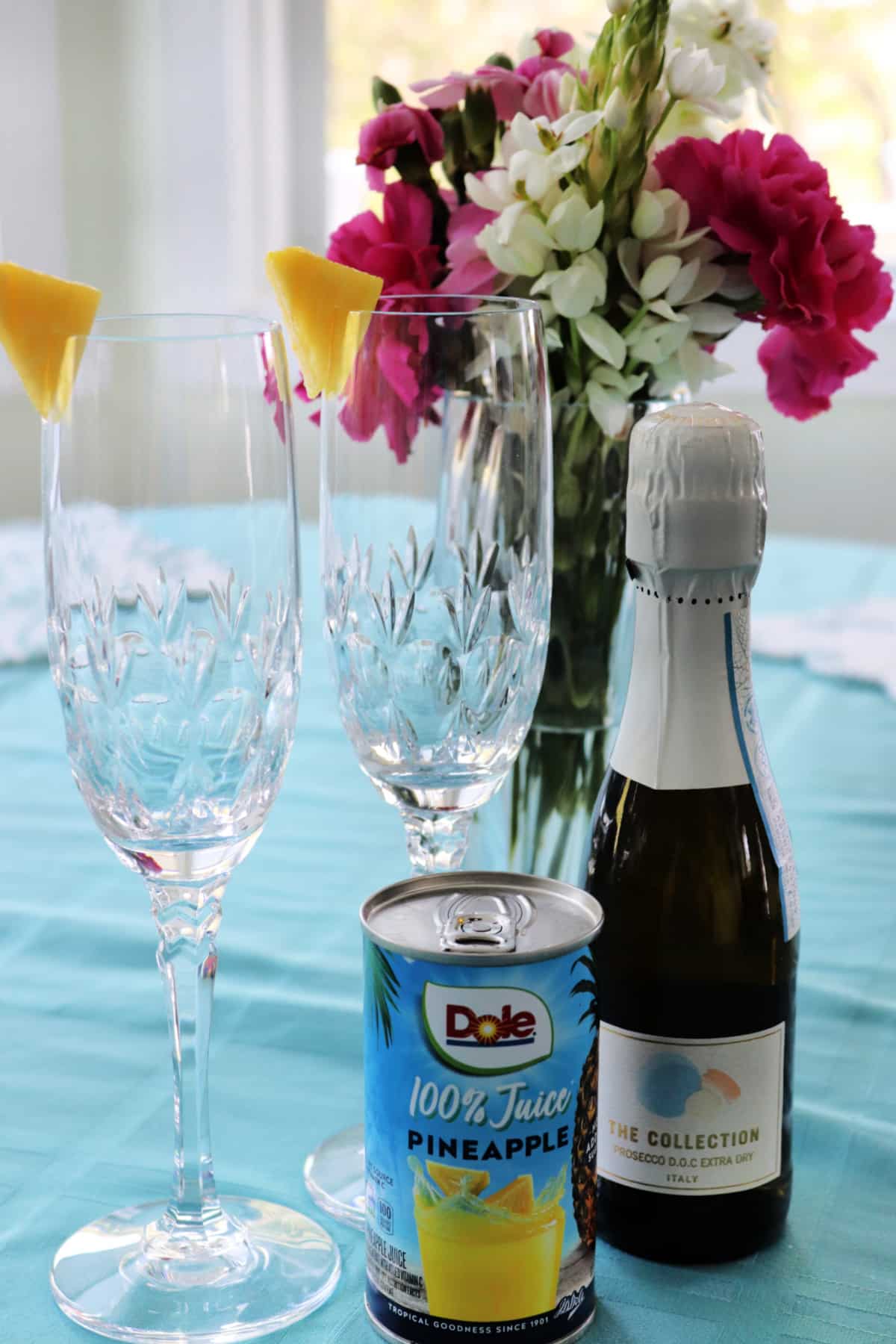 Two champagne glasses garnished with a pineapple wedge next to a small bottle of prosecco and a can of pineapple juice with a vase of flowers in the background.
