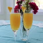 Two champagne glasses on a table with flowers behind them.