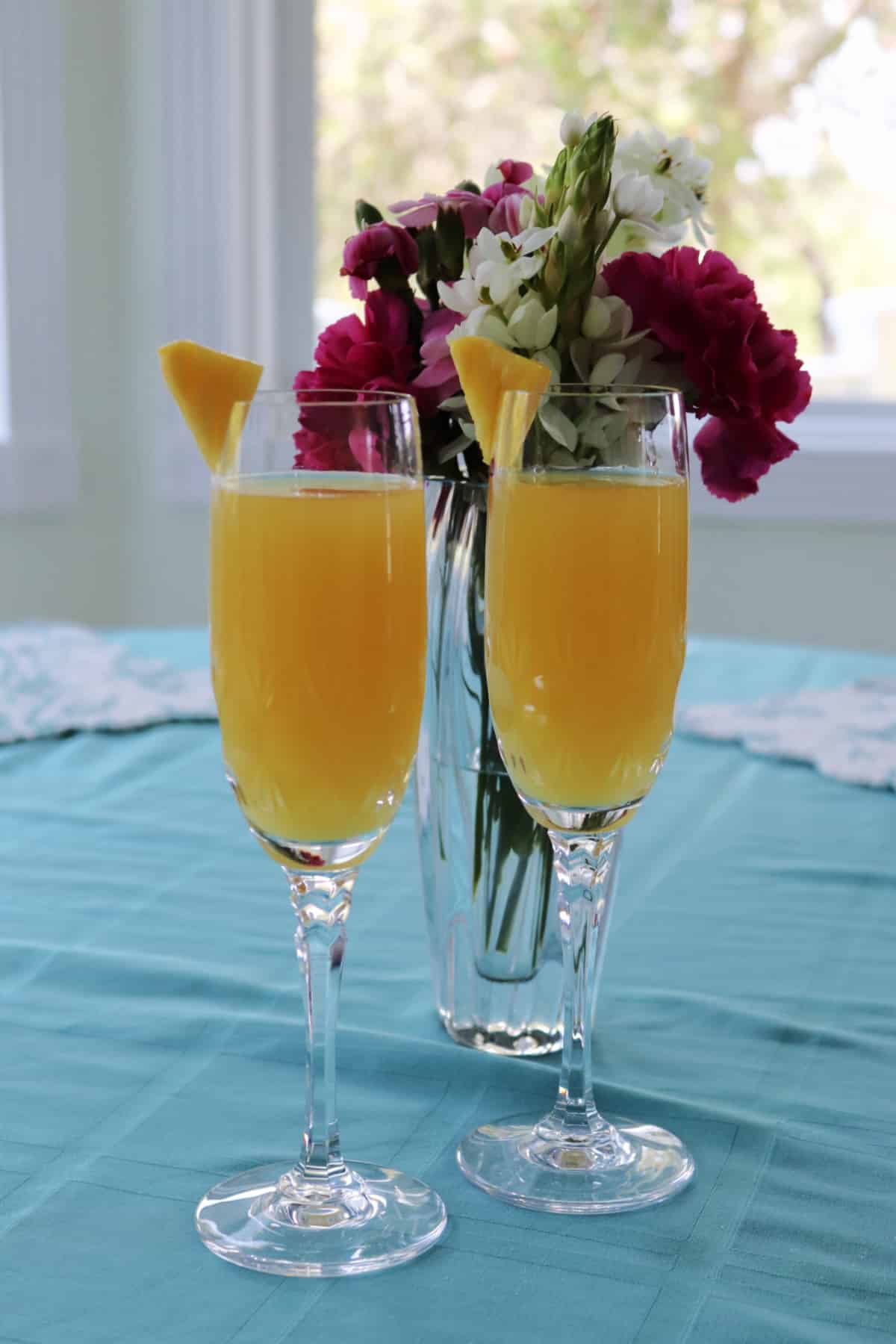 Two pineapple mimosa champagne glasses on a table with flowers behind them.