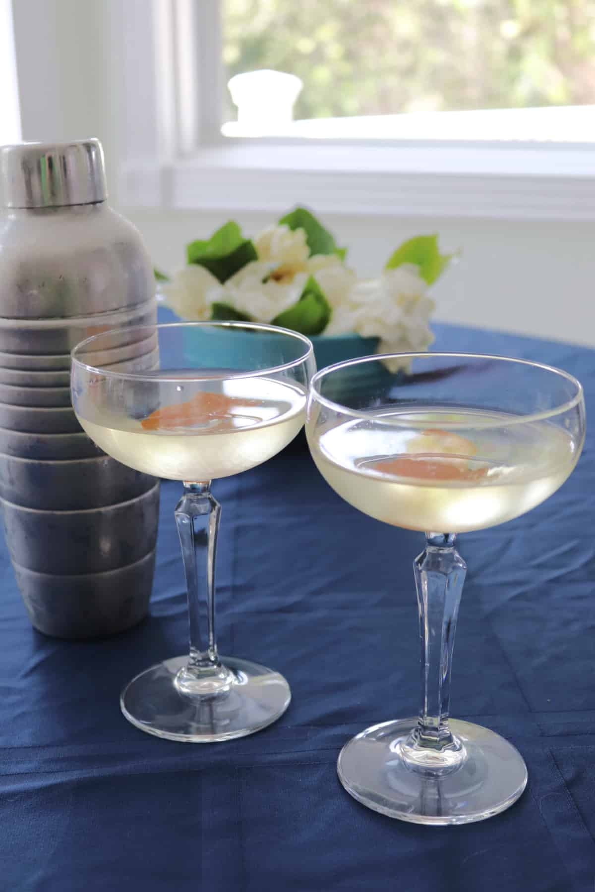 Two glasses of Saketini with a cocktail shaker next to them on a blue cloth.