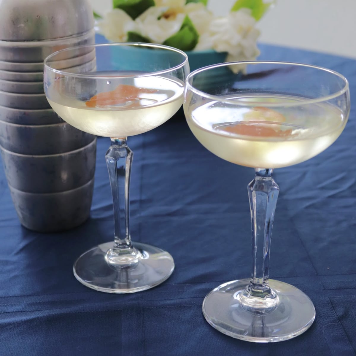 Two coupe glasses filled with liquid next to a shaker on a blue cloth
